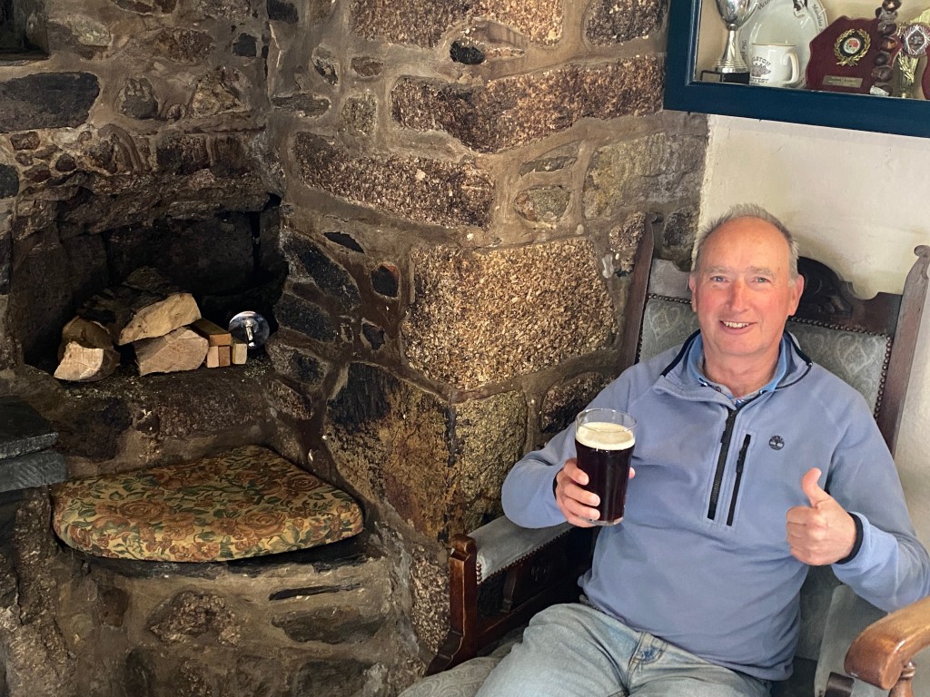 A man sitting in a cozy room next to a stone fireplace, smiling and giving a thumbs up while holding a pint of dark beer.