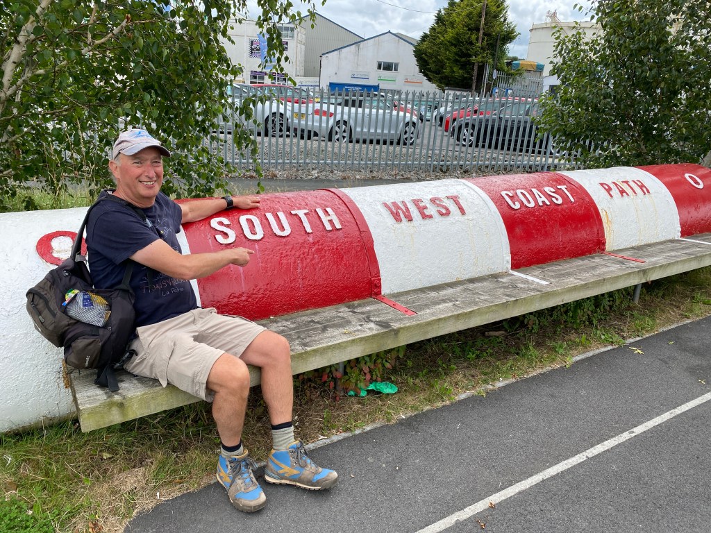 A man sitting on a wooden bench next to a large red and white sign that reads 'SOUTH WEST COAST PATH'. The setting features a few trees in the background and parked cars.