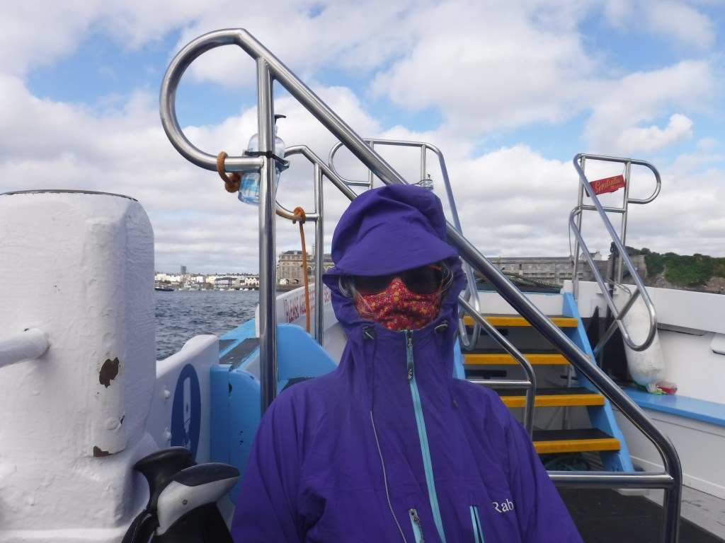 A person wearing a purple jacket, colorful mask, and sunglasses sits on a boat with a view of the water and distant buildings, under a partly cloudy sky.