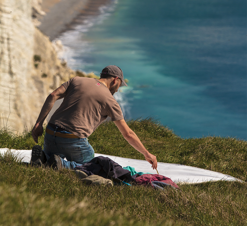 A person preparing a surfboard on a grassy hillside overlooking the ocean, with cliffs in the background.