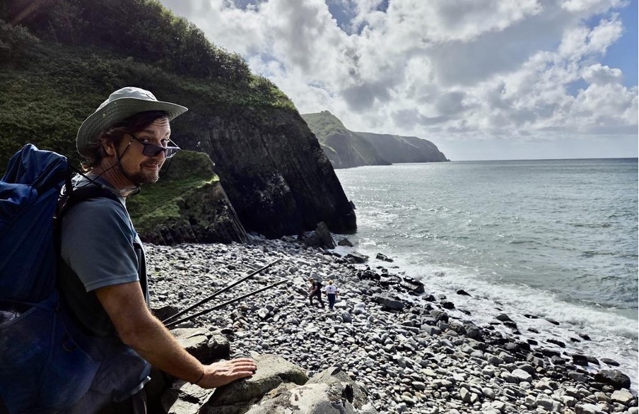 A man with a backpack and a hat stands on a rocky beach, looking towards the ocean and cliffs in the background. Two other individuals can be seen in the distance on the beach.
