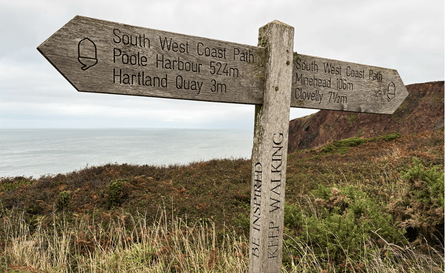 Wooden signpost indicating walking directions on the South West Coast Path, showing distances to Poole Harbour, Hartland Quay, Minehead, and Clovelly, with a scenic coastal background.