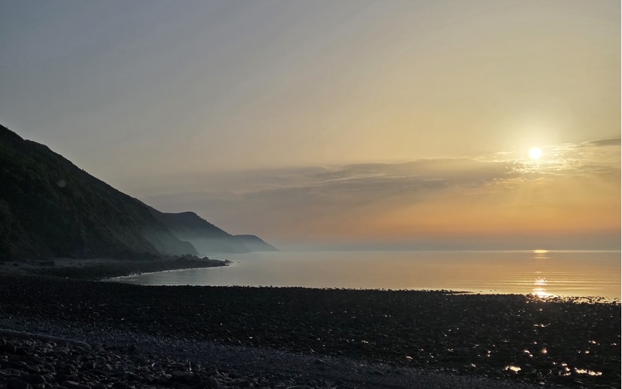 A serene coastal landscape at sunrise, featuring rolling hills and a calm sea reflecting warm hues in the sky.