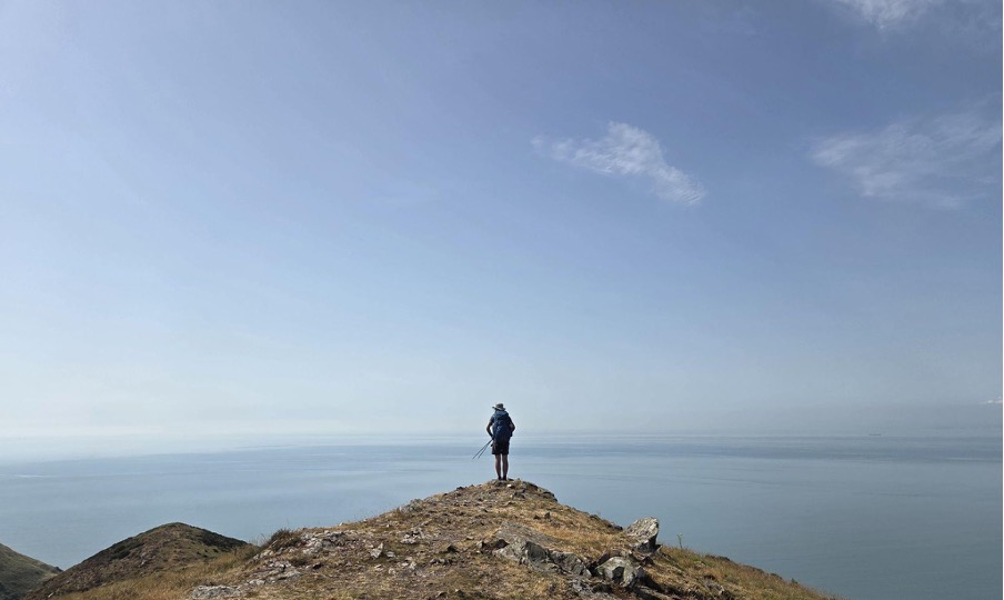 A hiker stands at the edge of a rocky cliff, overlooking a calm sea under a clear blue sky.