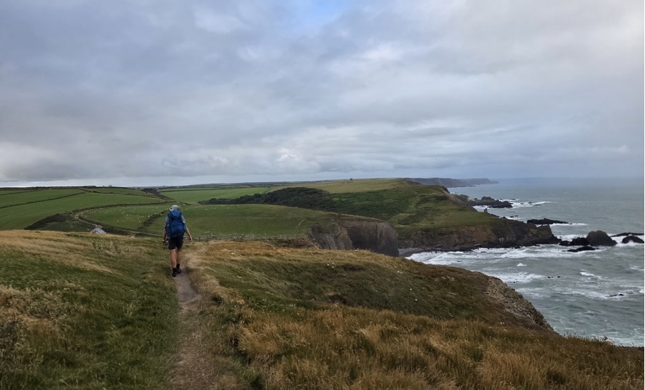 A hiker walking on a path along a coastal cliff with green fields and a cloudy sky in the background.